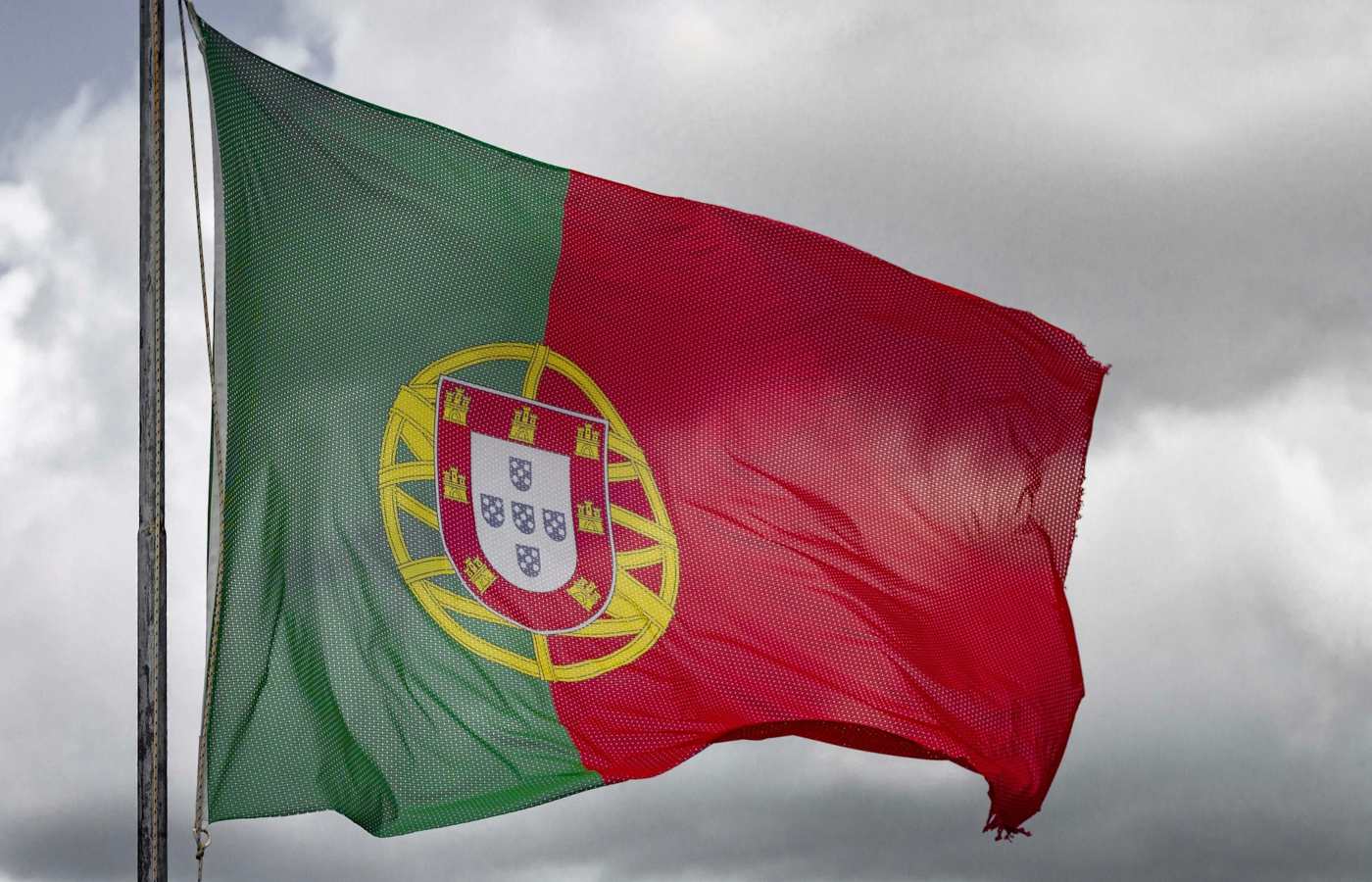 Bandeira de Portugal hasteada ao vento com céu nublado no fundo.